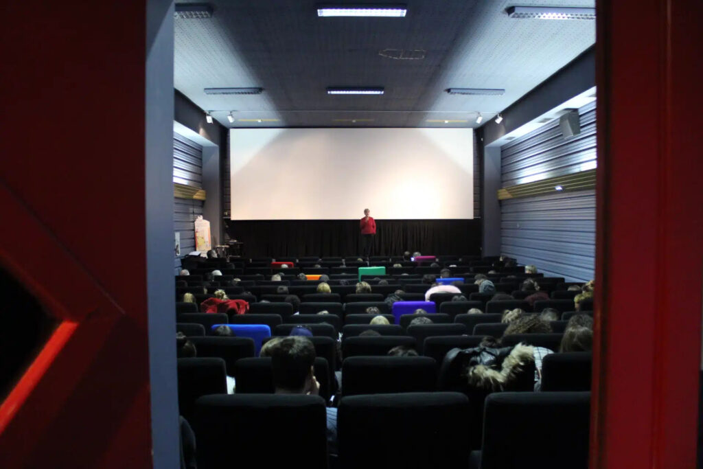 Salle de cinéma Le Rio avant le lancement d’un film à Clermont-Ferrand. La salle est presque pleine et une dame parle au micro sur le devant de la scène. Certains sièges sont colorés mais la majorité sont des sièges noirs.