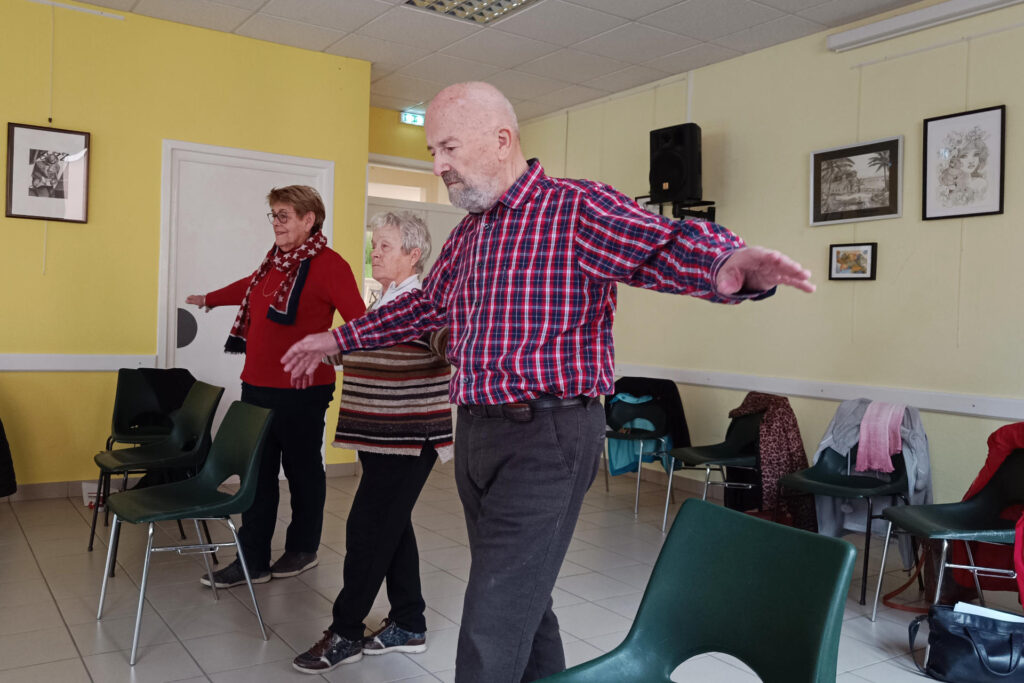 Sur cette photo, on voit trois personnes de plus de 60 ans. Il y a deux femmes, et un homme. Elles sont en train de réaliser un exercice durant l’atelier prévention des chutes. On les voit lever leurs deux bras horizontalement en mettant un pied devant l’autre. Proche d’elles, il y a des chaises pour s’appuyer en cas de perte d’équilibre.
