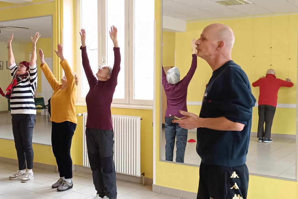 Sur cette photo, on voit trois femmes de plus de 60 ans et l’encadrant Arnaud. Elles sont en train de réaliser un exercice durant l’atelier prévention des chutes. On voit les trois femmes lever leurs deux bras vers le ciel. Le catch regarde un autre groupe. Dans le miroir, on voit le reflet de la femme (à droite sur la photo), et celui d’un participant masculin, en train de faire des flexions de bras contre un mur jaune.