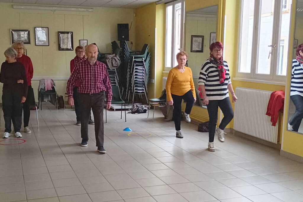 Sur cette photo, on voit six personnes de plus de 60 ans. Il y a cinq femmes et un homme. Il y a trois colonnes de deux personnes. Elles sont en train de réaliser un exercice durant l’atelier prévention des chutes. Elles montent un genou en marchant. Il y a un plot bleu et un cerceau jaune entre deux colonnes.