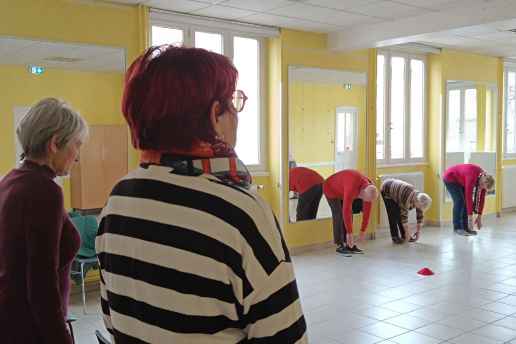 Sur cette photo, on voit cinq personnes de plus de 60 ans. Elles sont en train de réaliser un exercice durant l’atelier prévention des chutes. On voit deux femmes au premier plan qui se tiennent bien droite. Au second et au dernier plan, on voit deux femmes et un homme en train d’essayer ou de toucher leurs pieds, avec leurs deux mains, en étant debout, penchés vers le bas. Il y a un plot rouge.