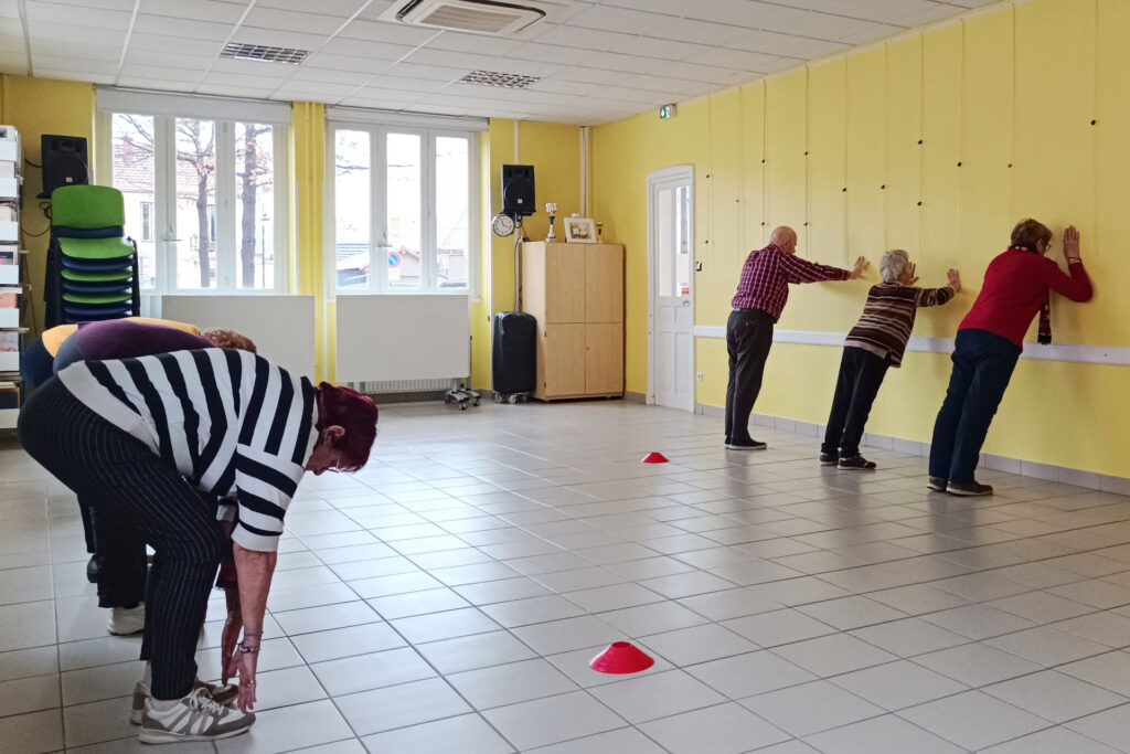 Sur cette photo, on voit six personnes de plus de 60 ans. Elles sont en train de réaliser un exercice durant l’atelier prévention des chutes. Il y a cinq femmes et un homme (deux des femmes ne sont presque pas visibles car cachées par la première). Trois personnes réalisent des flexions de bras contre le mur jaune. Trois autres personnes essayent de toucher leurs pieds, avec leurs deux mains, en étant debout et penchés vers le bas. Il y a deux plots rouges.