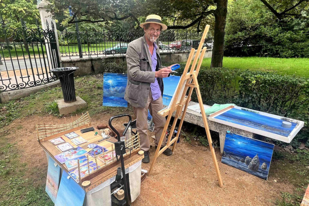 Sur cette photo, on voit le peintre Franck Martin, cigarette à la bouche, pinceau à la main. Devant lui, se tient son chevalet. À droite de l’image, on voit quelques une de ses œuvres, peintes dans des nuances de bleu, posées sur/contre le banc. Il y en a deux autres derrière lui. À gauche de l’image, sa petite table sur roulette lui servant à exposer ses autres œuvres. Franck Martin se trouve dans le cadre verdoyant du Parc des Sources.