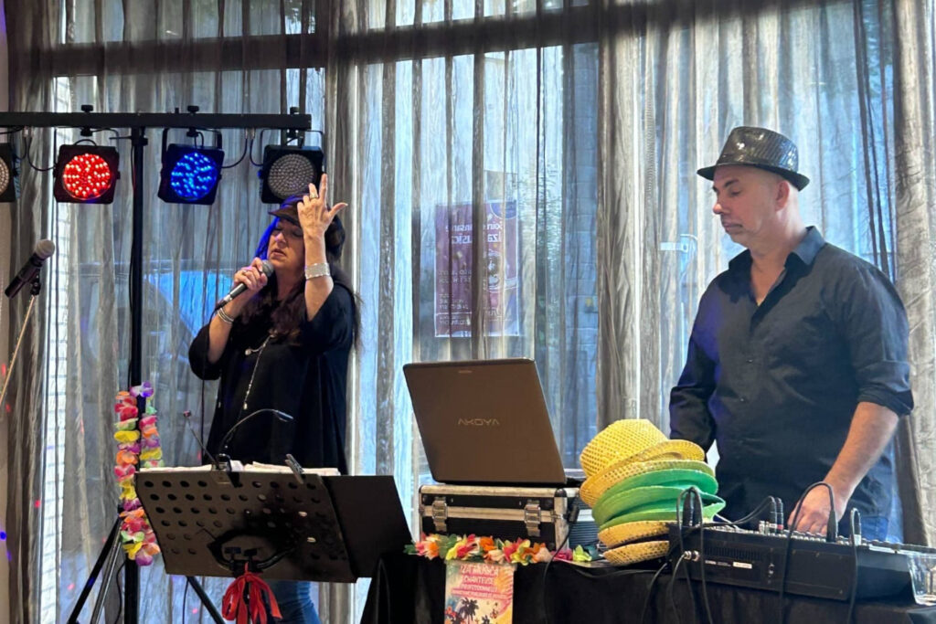 Il y a une scène animée d’un événement musical en intérieur. À gauche, une femme chante au micro (Iza Musica), semblant très impliquée dans sa performance, tandis qu’à droite, un homme, coiffé d’un chapeau noir, gère la musique depuis un ordinateur portable et une table de mixage. La pièce est éclairée par des lumières colorées projetant des points rouges, bleus et violets au plafond, créant une ambiance festive. Derrière eux, de grandes fenêtres recouvertes de rideaux laissent filtrer la lumière du jour. L’ensemble dégage une atmosphère conviviale et joyeuse.