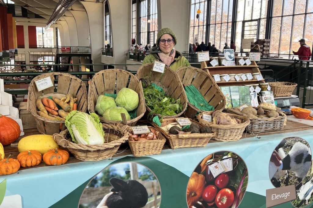 Sur cette photo, on voit la productrice Vicky Rohaut, vêtue d’un manteau et d’un bonnet vert, derrière son étal coloré. Celui-ci est rempli de paniers dans lesquels on retrouve des légumes de saison (carottes, courges, choux, échalottes, radis noirs, céleris, pommes de terres). L'arrière-plan montre les grandes fenêtres de la halle du Grand Marché. Son stand présente des images d'élevage et de maraîchage. Vicky porte des lunettes bleues.