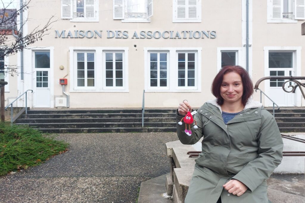 Au premier plan, Sophie Mestre est assise et tient un porte clef "goutte de sang". A l'arrière plan, la façade de la maison des associations de Saint-Yorre est reconnaissable.