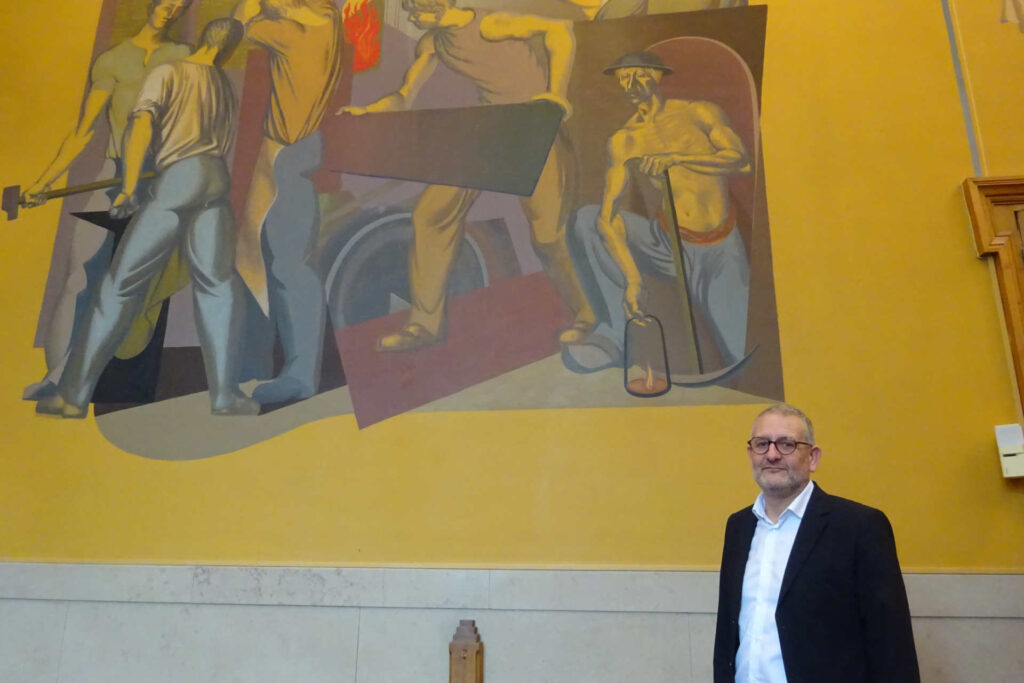 Un homme, ici, Sylvain Bourdier, le maire (DVG) de Commentry. Il pose devant une fresque ouvrière se trouvant dans la salle du conseil municipal.