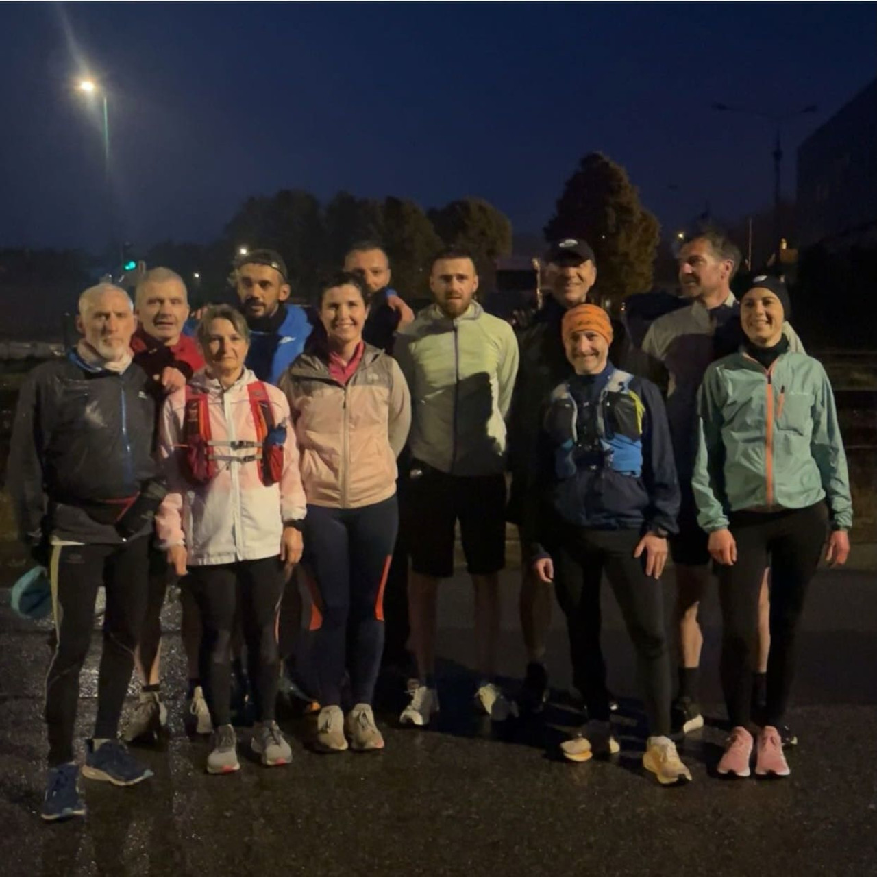 Un groupe de onze coureurs pose ensemble en extérieur, en tenue de sport, sur une route humide en soirée. Ils sont alignés, souriants, certains portant des sacs d’hydratation.