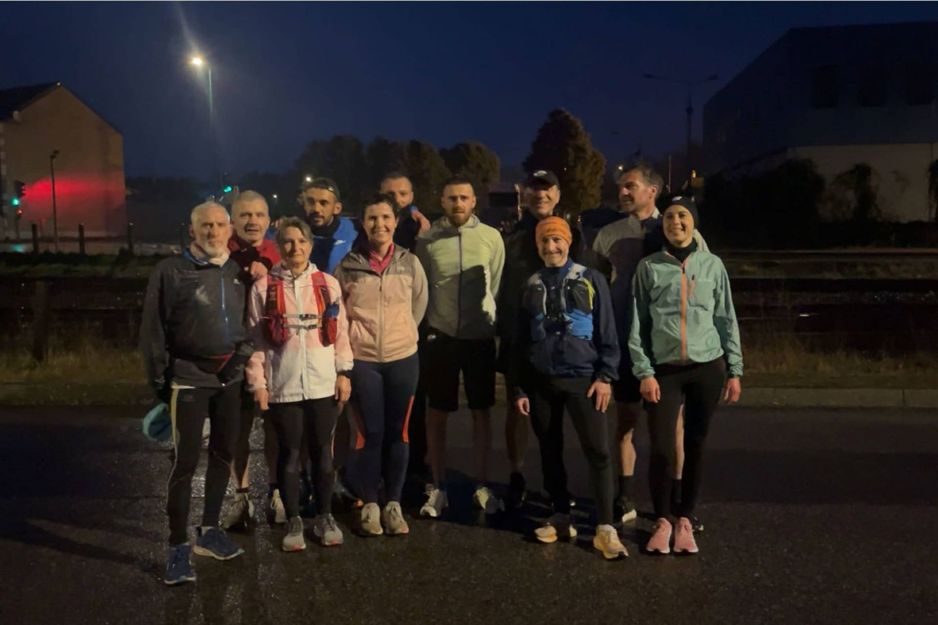 Un groupe de onze coureurs pose ensemble en extérieur, en tenue de sport, sur une route humide en soirée. Ils sont alignés, souriants, certains portant des sacs d’hydratation.