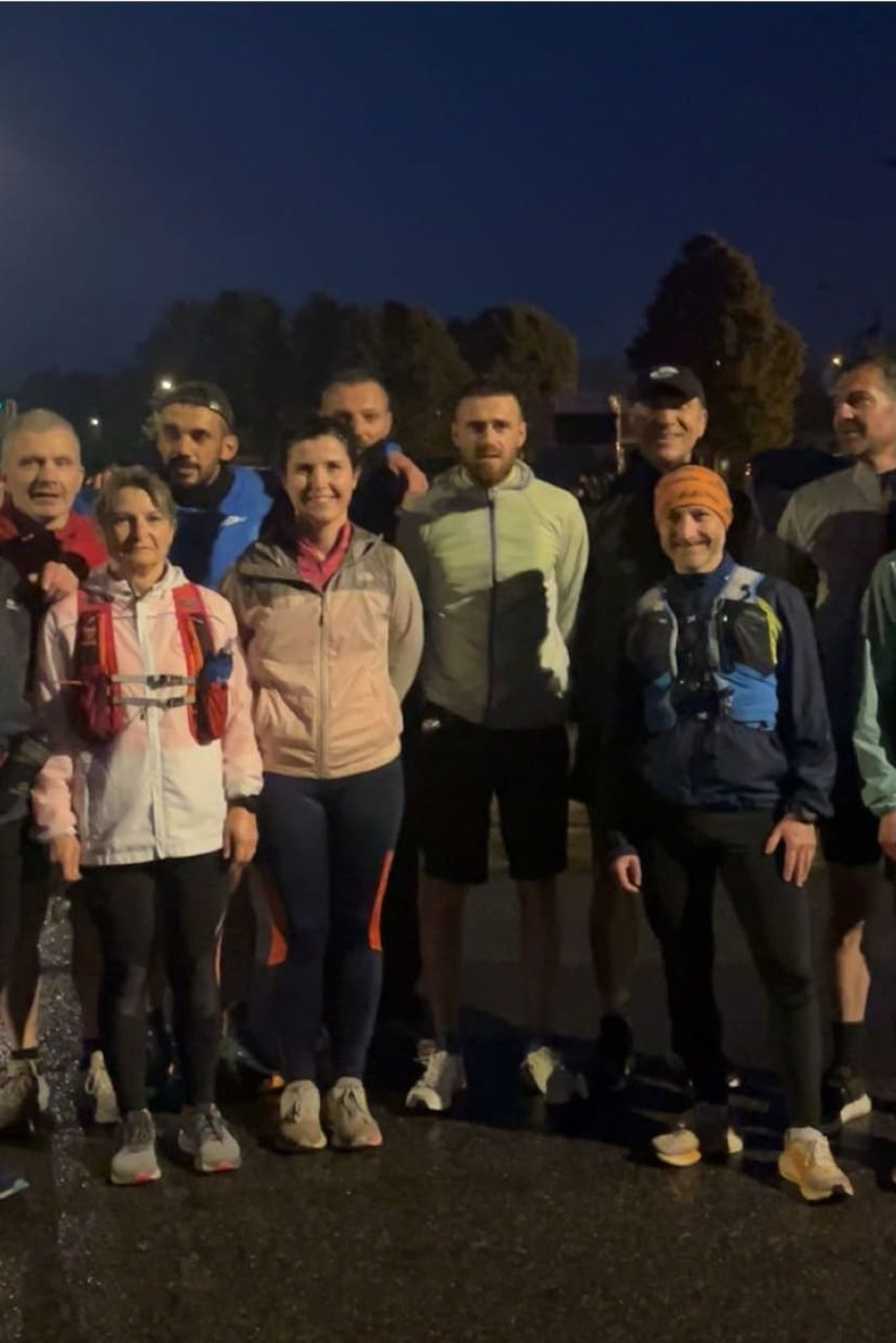 Un groupe de onze coureurs pose ensemble en extérieur, en tenue de sport, sur une route humide en soirée. Ils sont alignés, souriants, certains portant des sacs d’hydratation.
