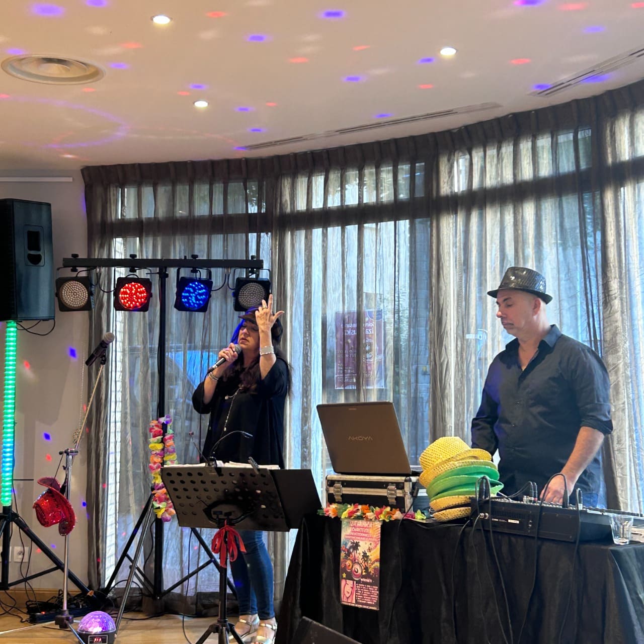 Il y a une scène animée d’un événement musical en intérieur. À gauche, une femme chante au micro (Iza Musica), semblant très impliquée dans sa performance, tandis qu’à droite, un homme, coiffé d’un chapeau noir, gère la musique depuis un ordinateur portable et une table de mixage. La pièce est éclairée par des lumières colorées projetant des points rouges, bleus et violets au plafond, créant une ambiance festive. Derrière eux, de grandes fenêtres recouvertes de rideaux laissent filtrer la lumière du jour. L’ensemble dégage une atmosphère conviviale et joyeuse.