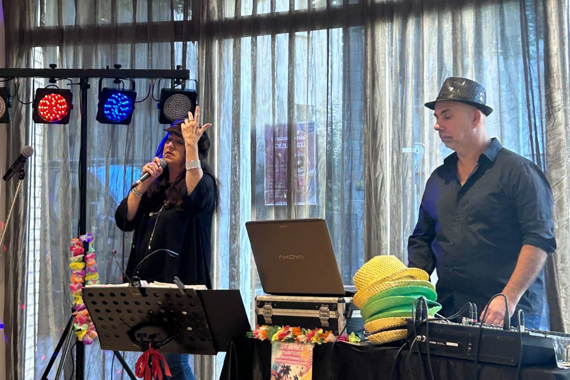 Il y a une scène animée d’un événement musical en intérieur. À gauche, une femme chante au micro (Iza Musica), semblant très impliquée dans sa performance, tandis qu’à droite, un homme, coiffé d’un chapeau noir, gère la musique depuis un ordinateur portable et une table de mixage. La pièce est éclairée par des lumières colorées projetant des points rouges, bleus et violets au plafond, créant une ambiance festive. Derrière eux, de grandes fenêtres recouvertes de rideaux laissent filtrer la lumière du jour. L’ensemble dégage une atmosphère conviviale et joyeuse.