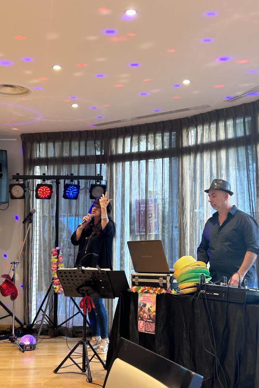 Il y a une scène animée d’un événement musical en intérieur. À gauche, une femme chante au micro (Iza Musica), semblant très impliquée dans sa performance, tandis qu’à droite, un homme, coiffé d’un chapeau noir, gère la musique depuis un ordinateur portable et une table de mixage. La pièce est éclairée par des lumières colorées projetant des points rouges, bleus et violets au plafond, créant une ambiance festive. Derrière eux, de grandes fenêtres recouvertes de rideaux laissent filtrer la lumière du jour. L’ensemble dégage une atmosphère conviviale et joyeuse.