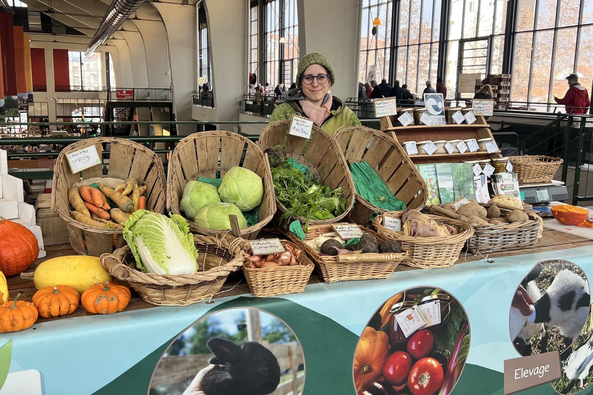 Sur cette photo, on voit la productrice Vicky Rohaut, vêtue d’un manteau et d’un bonnet vert, derrière son étal coloré. Celui-ci est rempli de paniers dans lesquels on retrouve des légumes de saison (carottes, courges, choux, échalottes, radis noirs, céleris, pommes de terres). L'arrière-plan montre les grandes fenêtres de la halle du Grand Marché. Son stand présente des images d'élevage et de maraîchage. Vicky porte des lunettes bleues.