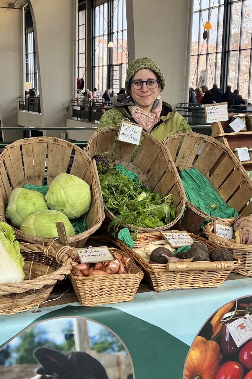 Sur cette photo, on voit la productrice Vicky Rohaut, vêtue d’un manteau et d’un bonnet vert, derrière son étal coloré. Celui-ci est rempli de paniers dans lesquels on retrouve des légumes de saison (carottes, courges, choux, échalottes, radis noirs, céleris, pommes de terres). L'arrière-plan montre les grandes fenêtres de la halle du Grand Marché. Son stand présente des images d'élevage et de maraîchage. Vicky porte des lunettes bleues.