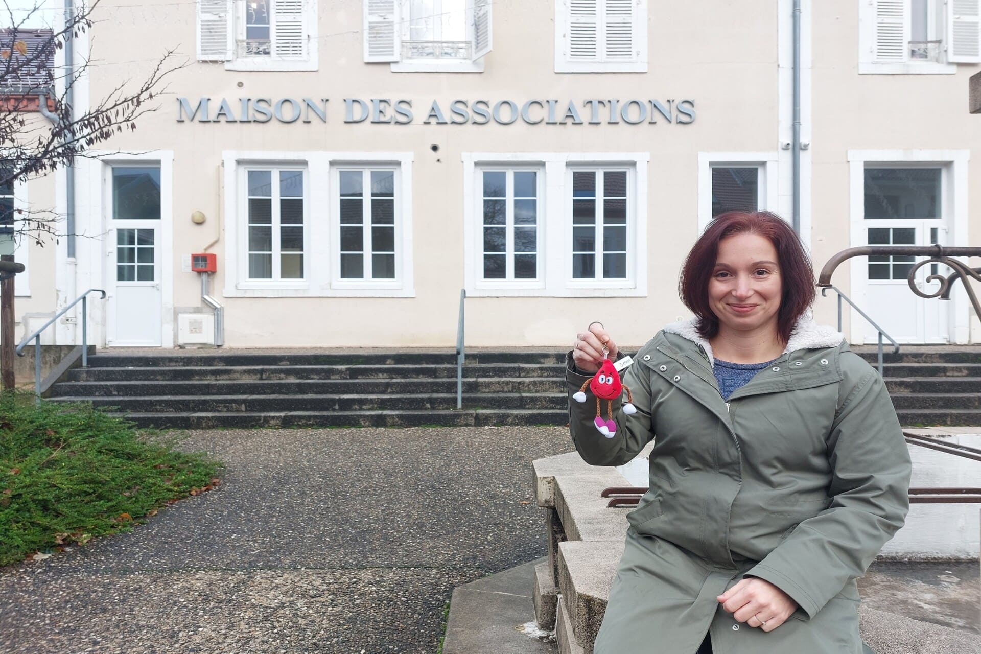 Au premier plan, Sophie Mestre est assise et tient un porte clef "goutte de sang". A l'arrière plan, la façade de la maison des associations de Saint-Yorre est reconnaissable.