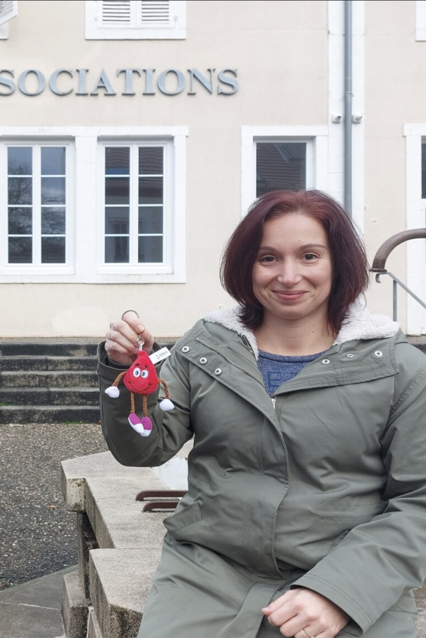 Au premier plan, Sophie Mestre est assise et tient un porte clef "goutte de sang". A l'arrière plan, la façade de la maison des associations de Saint-Yorre est reconnaissable.